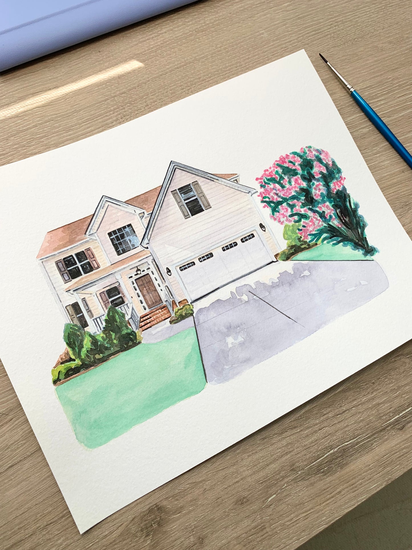 watercolor home portrait on a wooden surface next to a paint brush