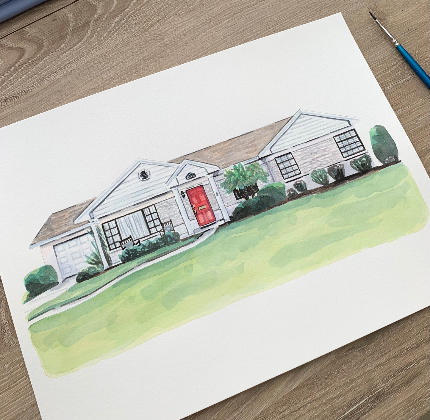Watercolor painting of a house on a wooden surface with a paint brush