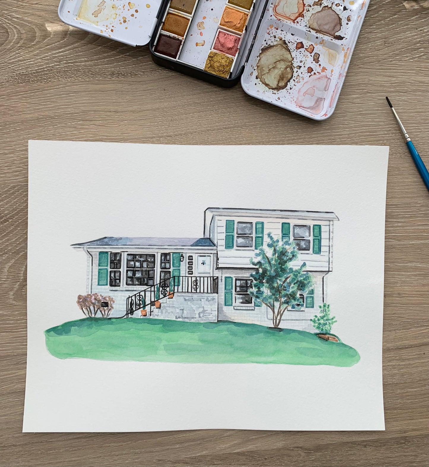 Watercolor painting of a house on a wooden surface with watercolor pans and a brush.