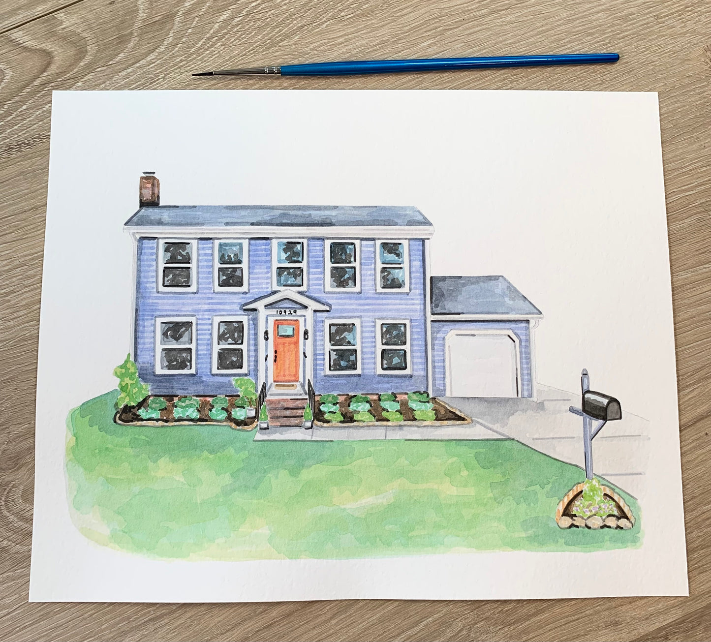 Watercolor painting of a house on a wooden surface with a paintbrush.