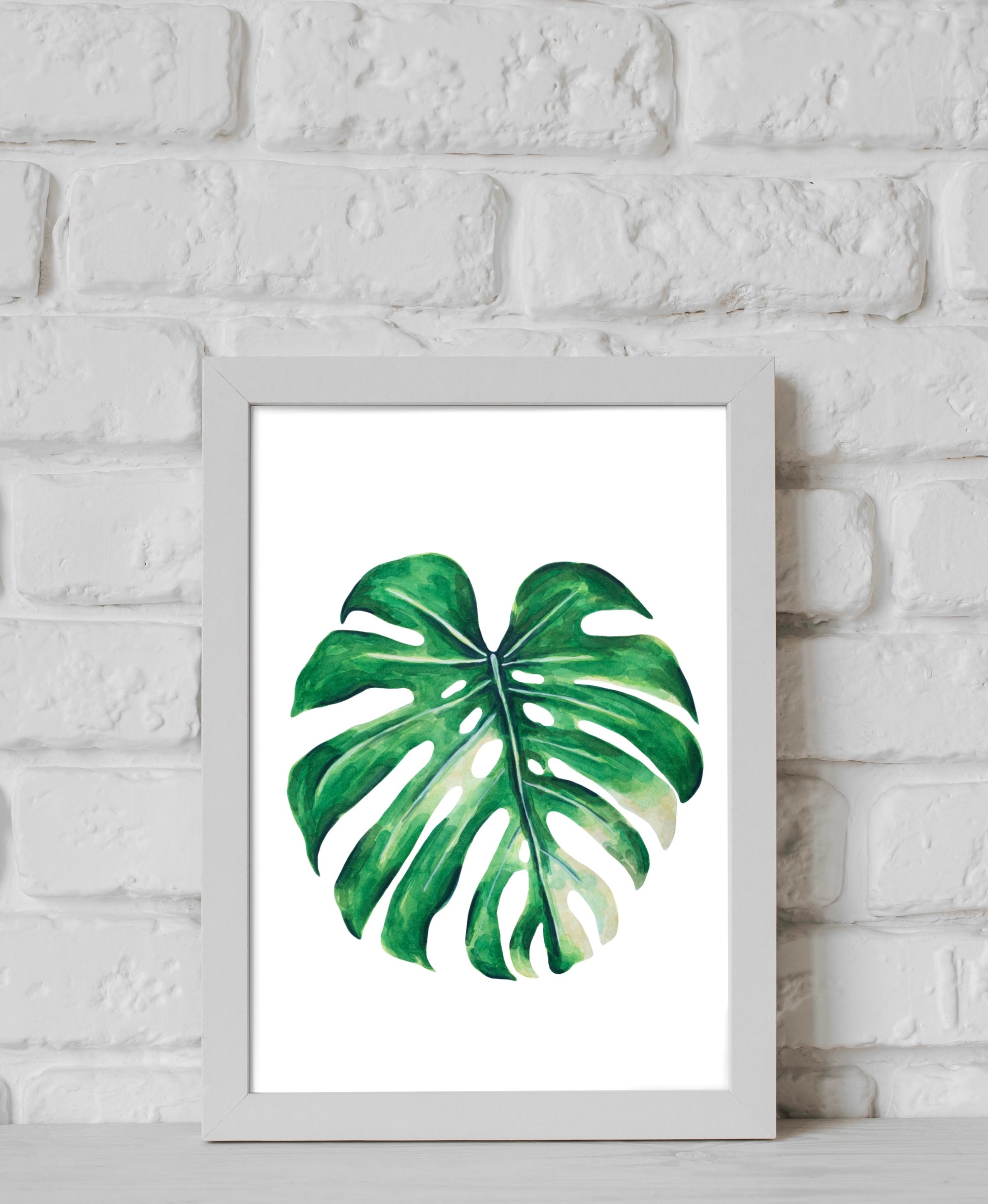 Framed artwork of a monstera leaf on a white brick wall background