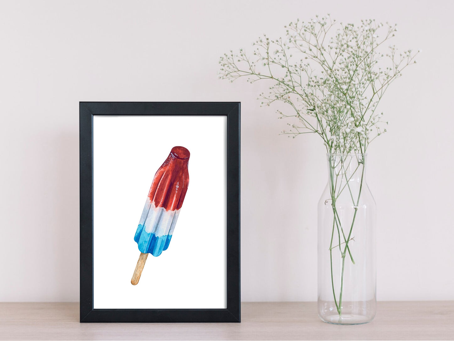 Framed artwork of a red, white, and blue popsicle on a white wall with a vase of flowers.
